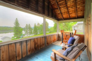 Eine überdachte Veranda mit Blick auf einen See und Bäume, Holzmöbel und ein Haus im Hintergrund.