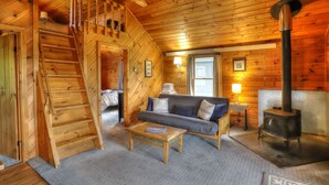 A cozy cabin interior with a wooden staircase, a sofa, a coffee table, and a wood-burning stove. - Hunter Cove Cabin #3: Shared Rangeley Lakefront, Dock Slip + Screened Porch (Rangeley)