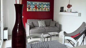 Una sala de estar moderna con una pared de acento roja, un sofá gris, un sillón blanco, un jarrón negro y un cuadro de casas coloridas.