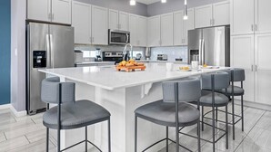 Cocina moderna con armarios blancos, electrodomésticos de acero inoxidable, una isla central con taburetes y luminarias colgantes.