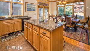 A kitchen with wooden cabinets, granite countertops, and a large island. There is a dining area with a table and chairs, and large windows offering a view of the outside. - Charming Big Sky Condo! Cozy Fireplace, 10 Min Drive to Resort, 3 Min drive to T (Big Sky)