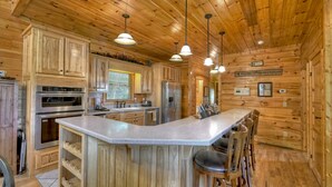 Cocina de cabaña de madera con una isla central, taburetes de bar, electrodomésticos de acero inoxidable y un letrero que dice 'Bienvenido a las Montañas'.