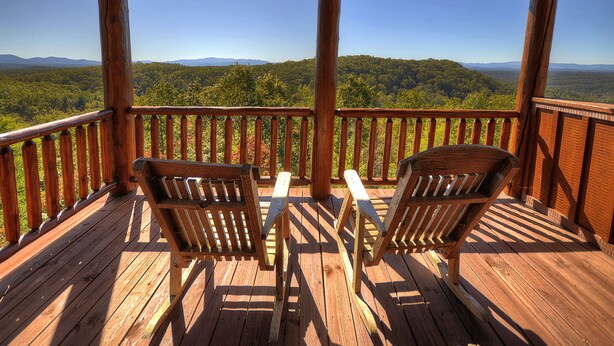 Una terraza de madera con dos sillas y una mesa, con vistas a un paisaje boscoso y montañas a lo lejos.