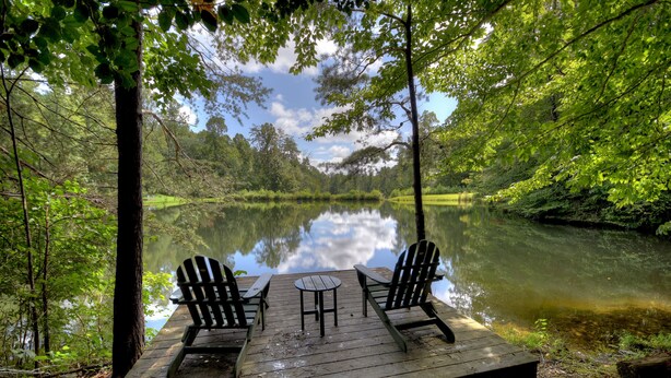 A wooden deck with two Adirondack chairs and a small table overlooking a calm lake, surrounded by lush greenery. - Pate`s Escape - Cherry Log (Cherry Log)