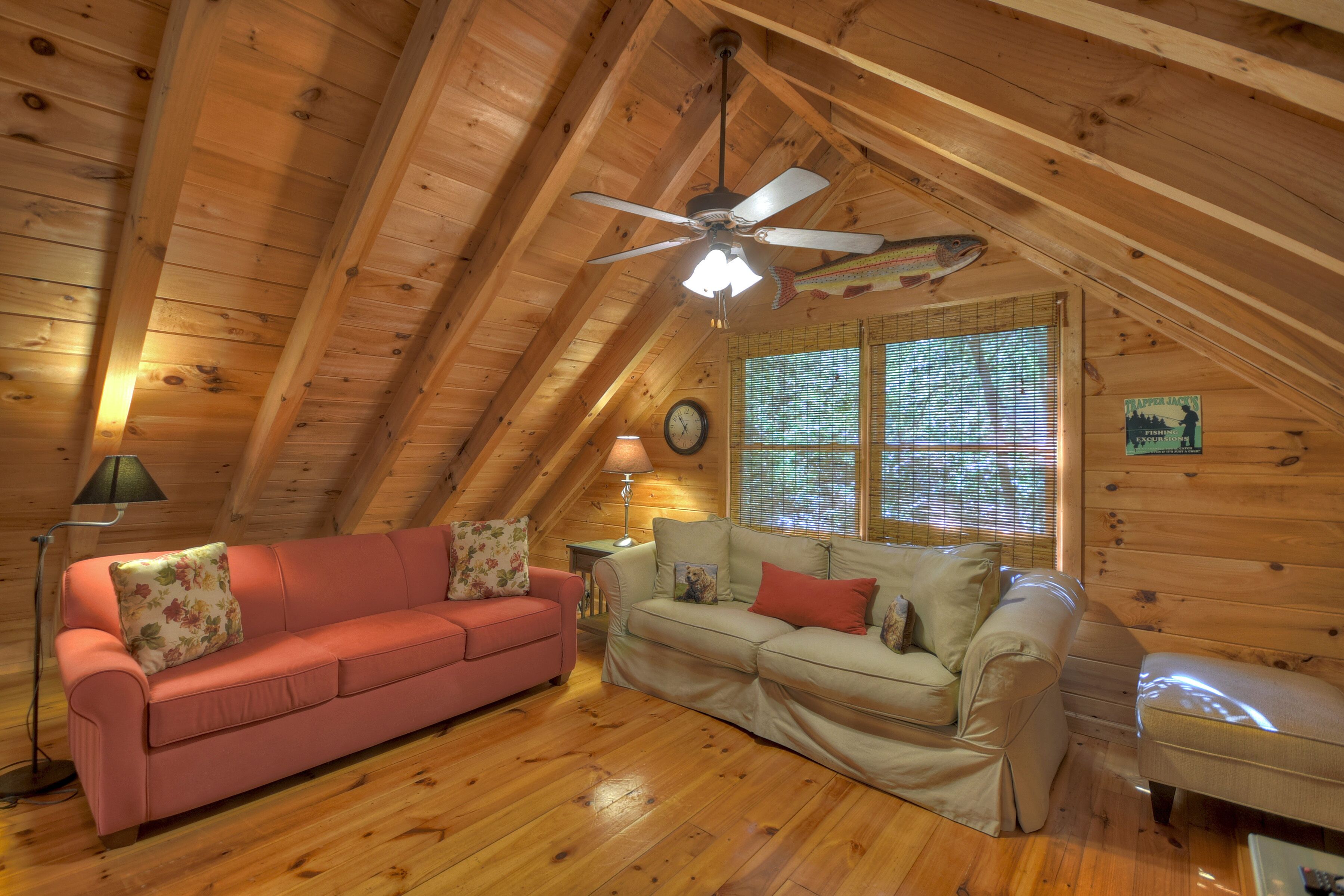 A cozy attic room with wooden walls and a sloped ceiling. There are two sofas, a lamp, and a clock on the wall.