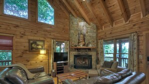A cozy cabin interior with a stone fireplace, leather sofas, a television, and large windows. - Pate`s Escape - Cherry Log (Cherry Log)
