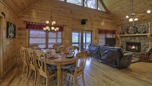 A wooden cabin interior with a dining table, chairs, a leather sofa, and a stone fireplace.