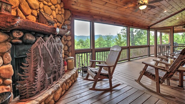 A rustic cabin with a stone fireplace, wooden rocking chairs, and large windows overlooking a forested landscape.