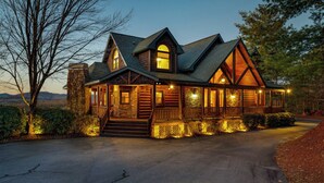 Une grande cabane en rondins avec une cheminée en pierre, de nombreuses fenêtres et une véranda décorée de guirlandes lumineuses.