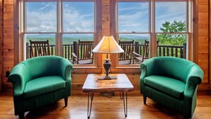 Une pièce aux murs en bois et aux grandes fenêtres, avec deux fauteuils verts, une petite table surmontée d’une lampe, et une vue sur une terrasse et le paysage environnant.