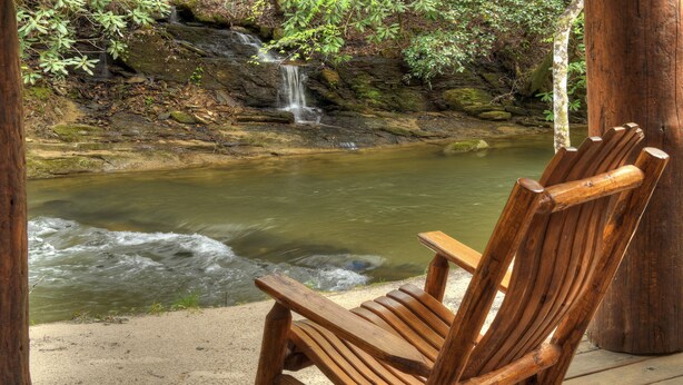 Ein Holzsessel blickt auf einen kleinen Wasserfall und einen Fluss, mit einem hölzernen Deck und einem Baumstamm in der Nähe.