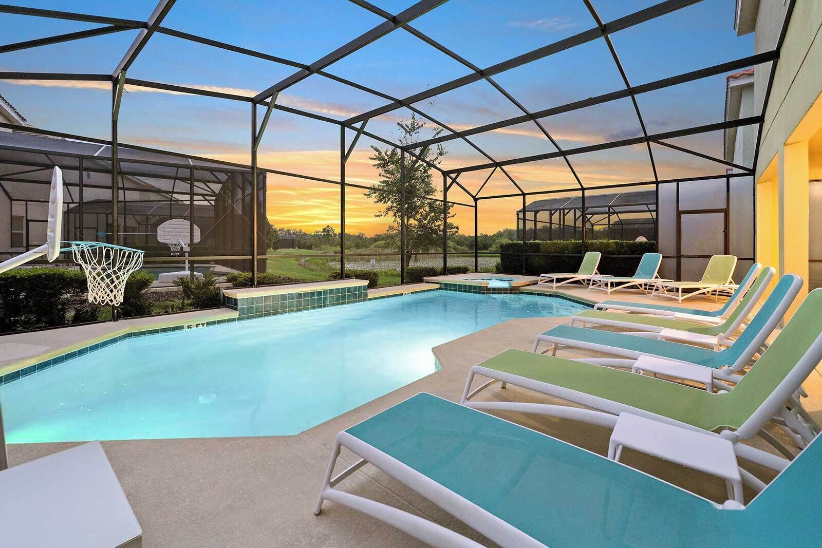 A pool area with a basketball hoop, lounge chairs, and a view of the sunset through a glass enclosure.