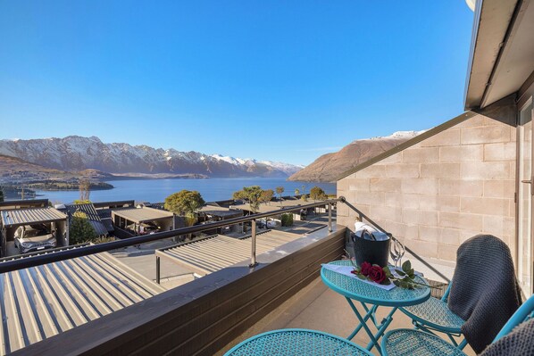 A balcony with a view of mountains and a body of water. There are two blue chairs and a table with a rose and a bucket.