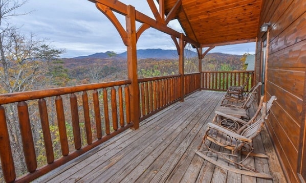 A wooden deck with a railing, rustic wooden chairs, and a view of autumn foliage and mountains.