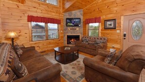 Interior acogedor de una cabaña de troncos con chimenea, paredes de madera, un televisor montado arriba y una zona de estar cómoda con dos sofás y una mesa de centro.