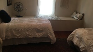 Ett sovrum med två sängar, ett fönster med tunna gardiner, ett element och en väggmonterad lampa.