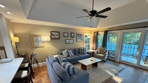 A modern living room with a grey sofa, a coffee table, and a ceiling fan. There are framed pictures on the wall and a view of the outdoors through large sliding doors.