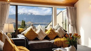 Ein gemütliches Wohnzimmer mit einer Couch, Kissen und einer Lampe. Ein großes Fenster bietet Blick auf Berge und einen See.