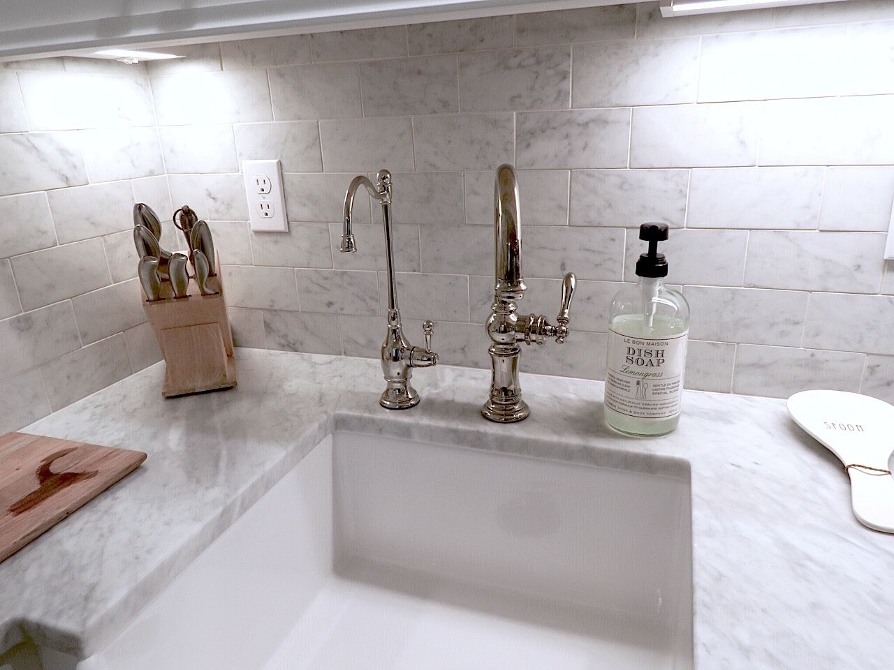 A bathroom with a white sink, two faucets, a soap dispenser, and a wooden cutting board.