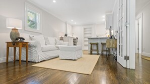 A spacious living room with a sofa, ottoman, and a small table with a lamp. There is a kitchen area with a bar stool and a wooden chair.
