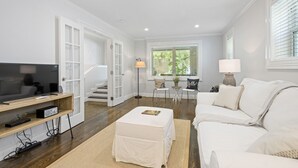 A modern living room with a flat-screen TV, a white sofa, a coffee table, and a dining area with two chairs and a small table.