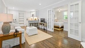 A spacious living room with a sofa, ottoman, and a coffee table. There is a kitchen area with a dining table and chairs. The room has a large window and a door leading to another room.