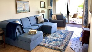 A living room with a blue sofa, a patterned rug, and a painting of a ship on the wall. - Best views on MDI - 2 bedroom 2 bath condo - waterfront with in ground pool (Southwest Harbor)
