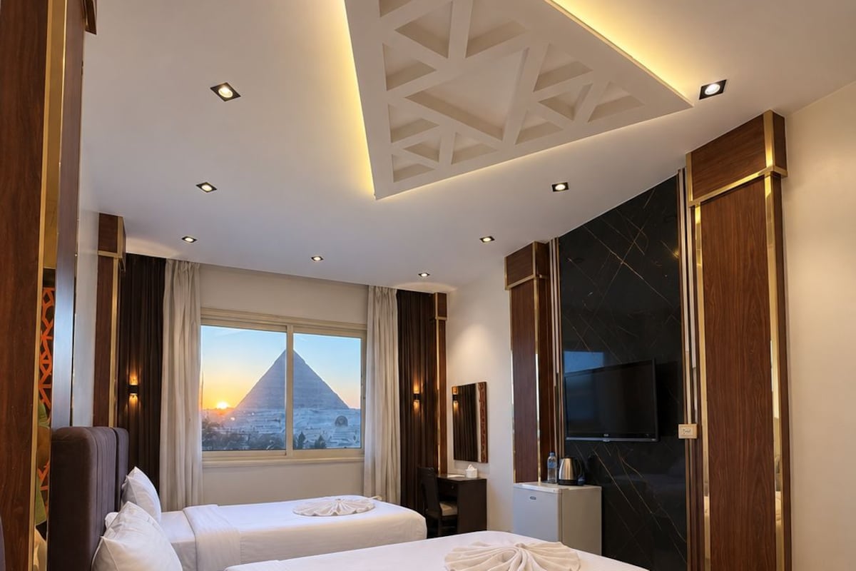 Quarto de hotel com duas camas, televisão, escrivaninha e vista para uma pirâmide.