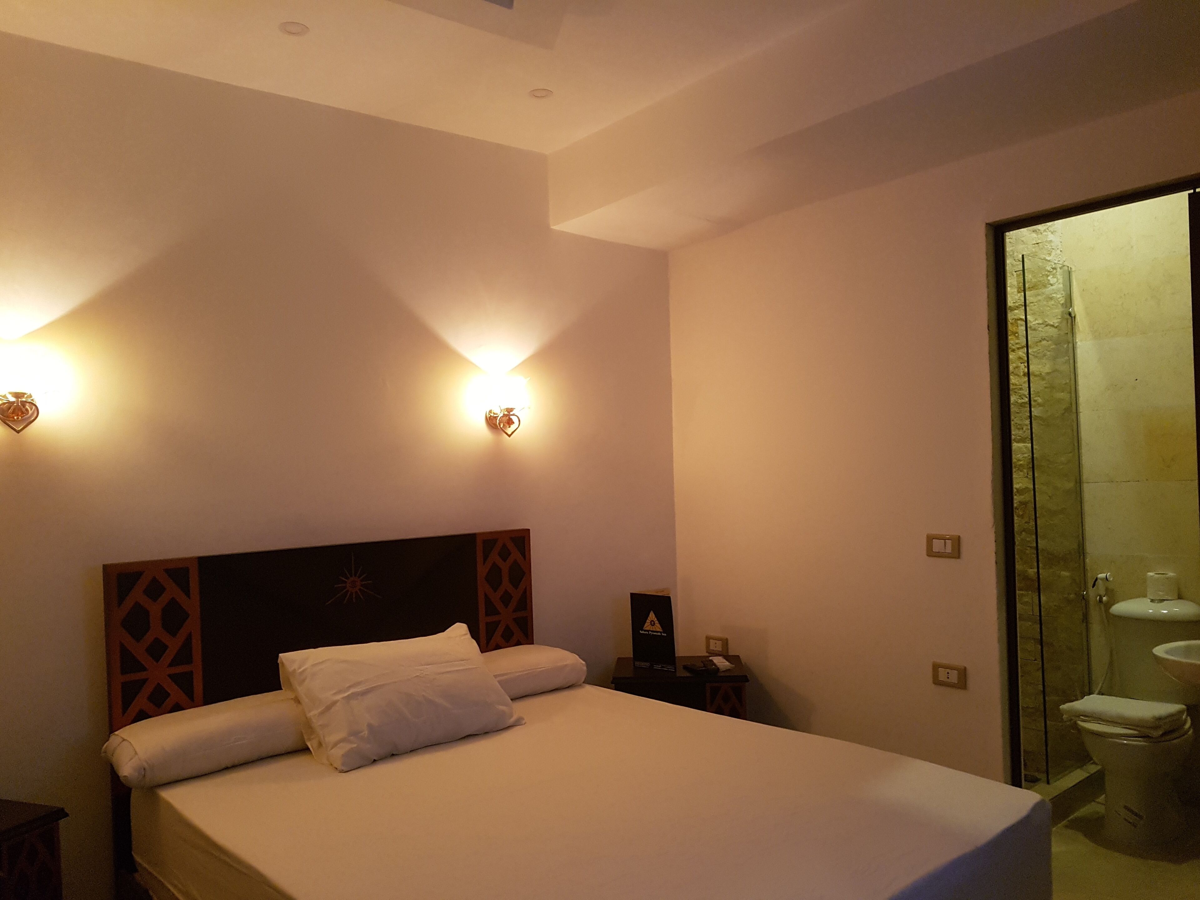 A hotel room with a bed, a nightstand, a wall-mounted light, and a bathroom with a toilet and shower.