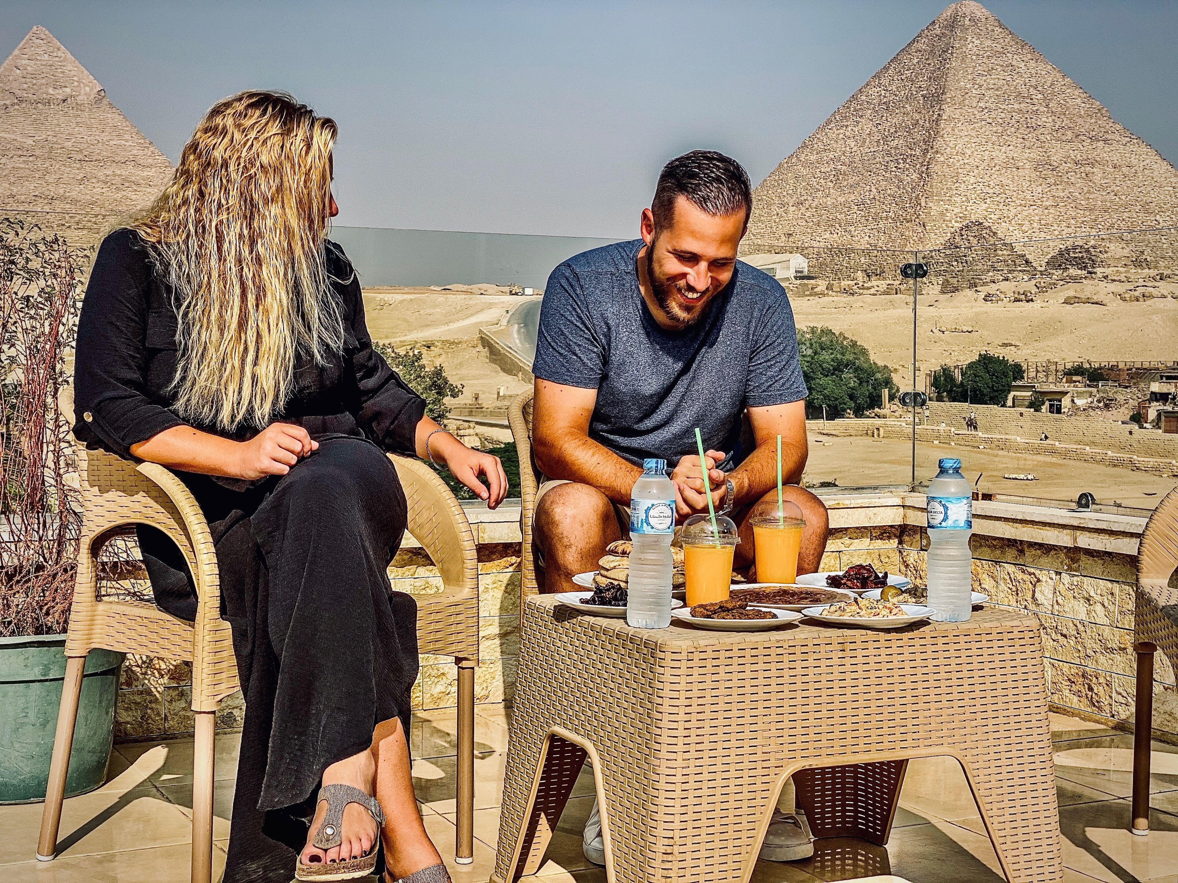 Zwei Personen sitzen an einem Tisch mit Getränken und Speisen, Pyramiden sind im Hintergrund zu sehen.