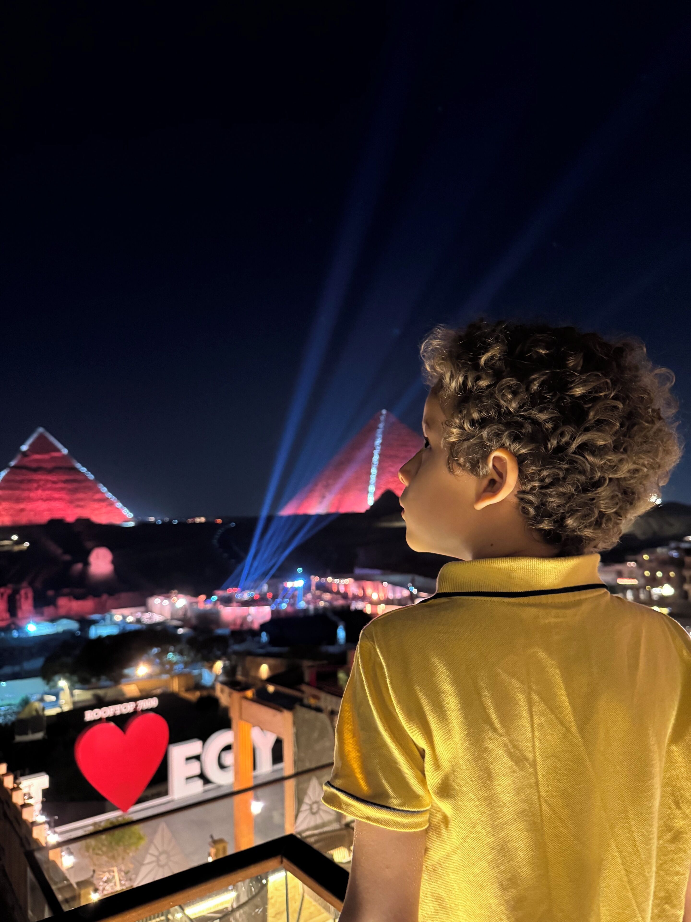 Una persona de pie en un balcón con vistas a un paisaje urbano donde se ven pirámides iluminadas y un edificio destacado con el letrero "EGYPT".