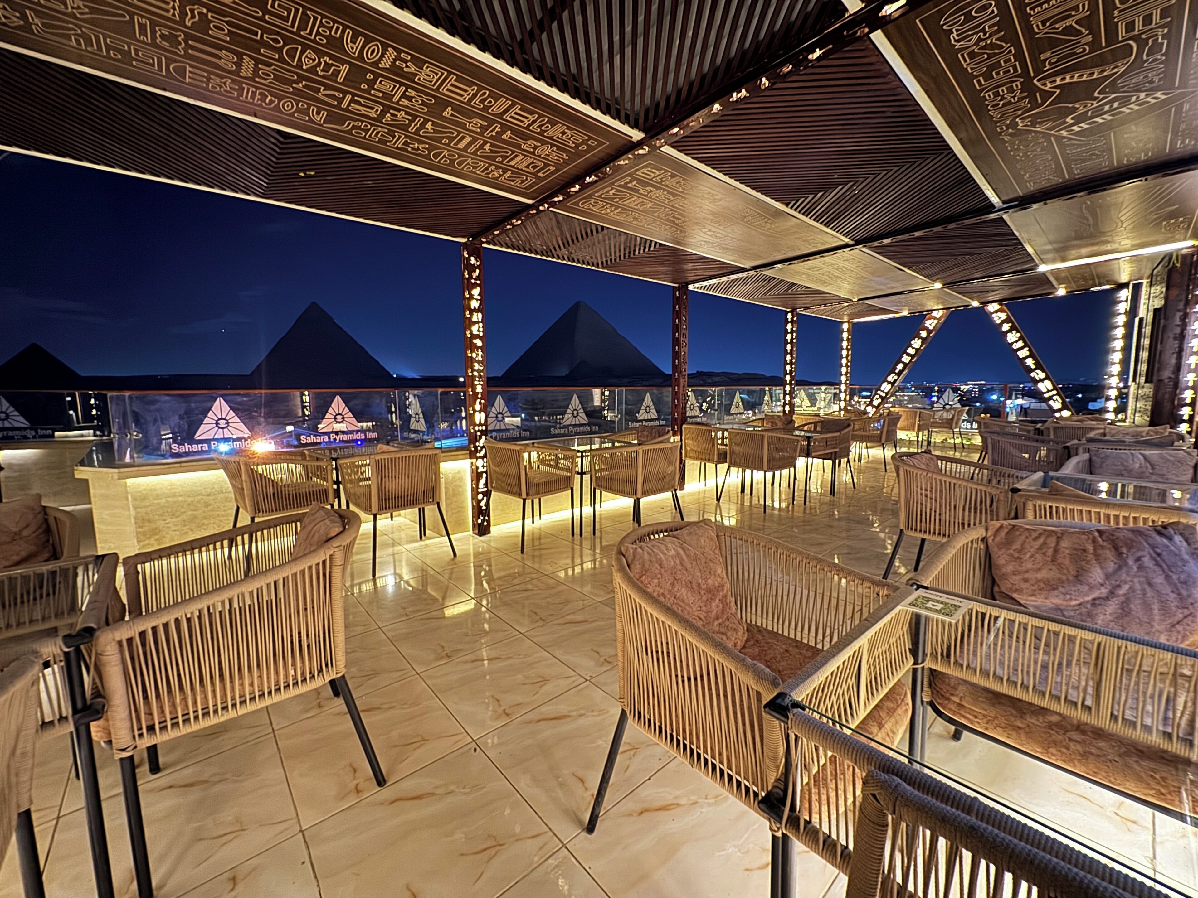 Un espace extérieur aménagé avec des chaises en osier et un bar, offrant une vue sur les pyramides illuminées la nuit.