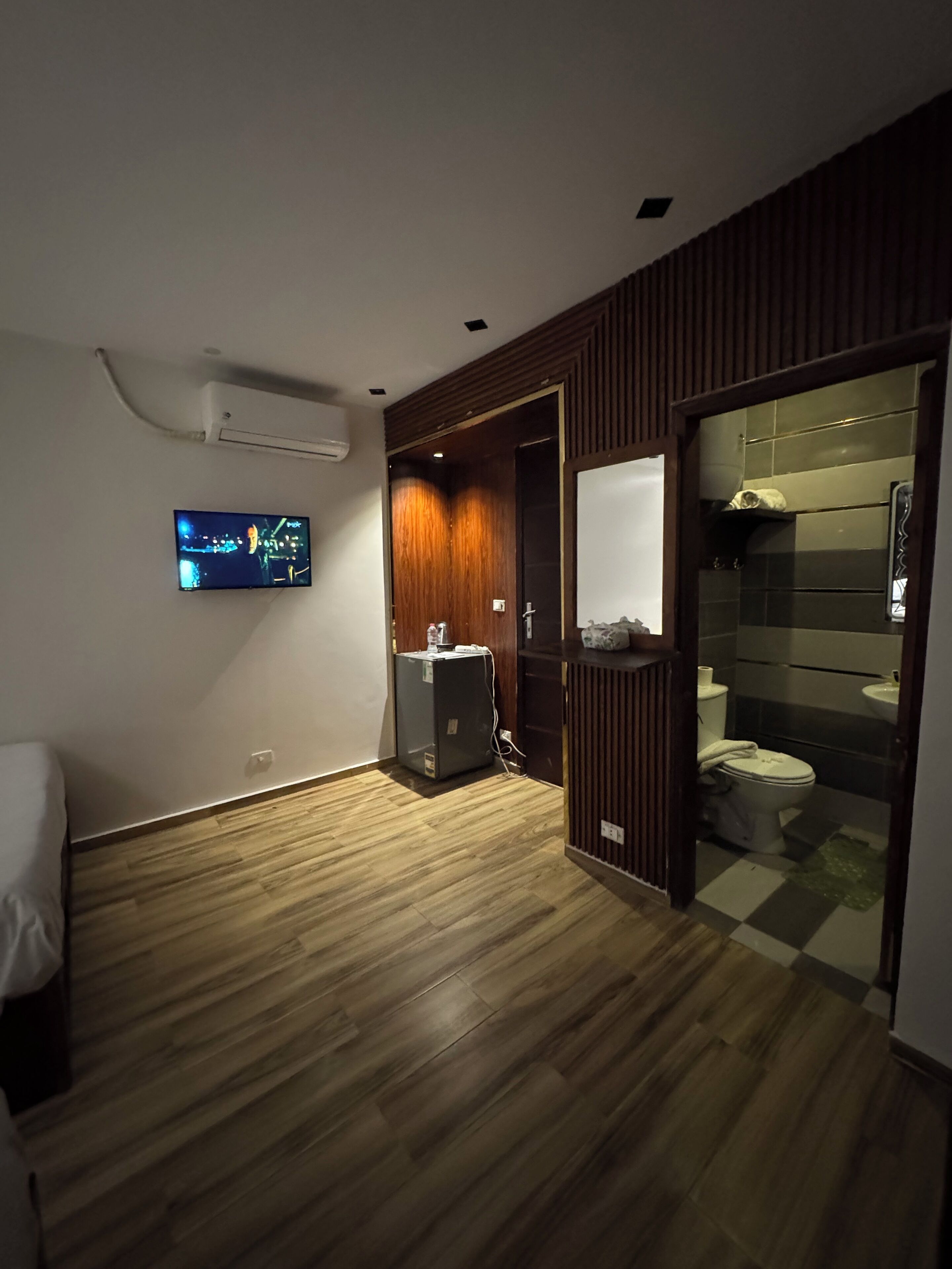 Ein Hotelzimmer mit Holzboden, einem Bett, einem an der Wand befestigten Fernseher, einem Kühlschrank und einem Badezimmer mit Schachbrettmuster.