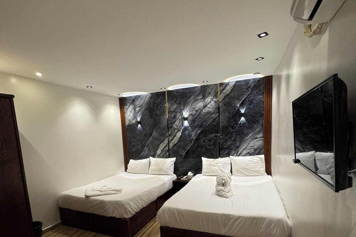 A modern hotel room with two single beds, wooden flooring, a marble accent wall, and a mounted TV.