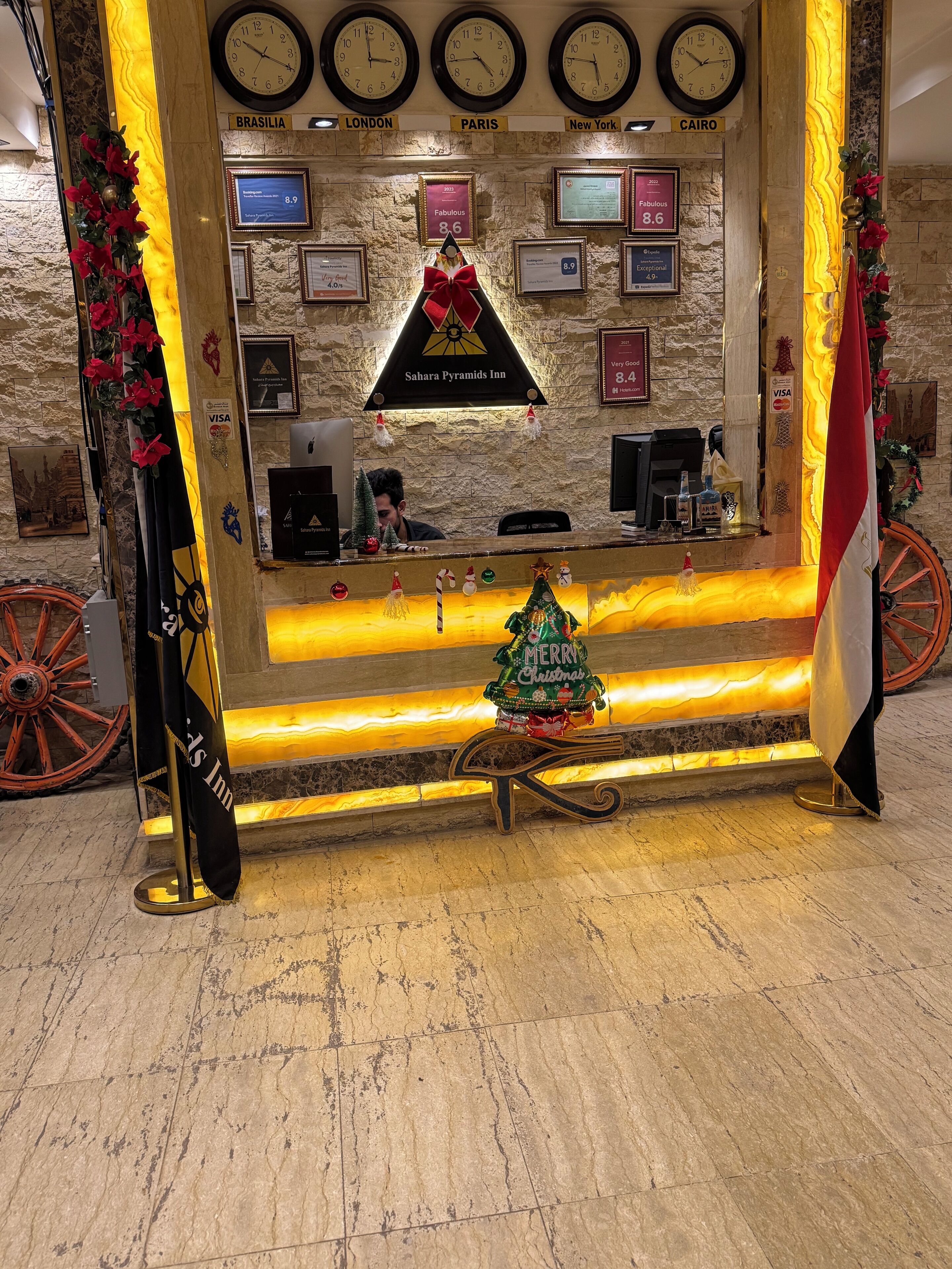 Zona de recepción con relojes que muestran diferentes ciudades, un árbol de Navidad y un letrero que indica el Sahara Pyramids Inn.