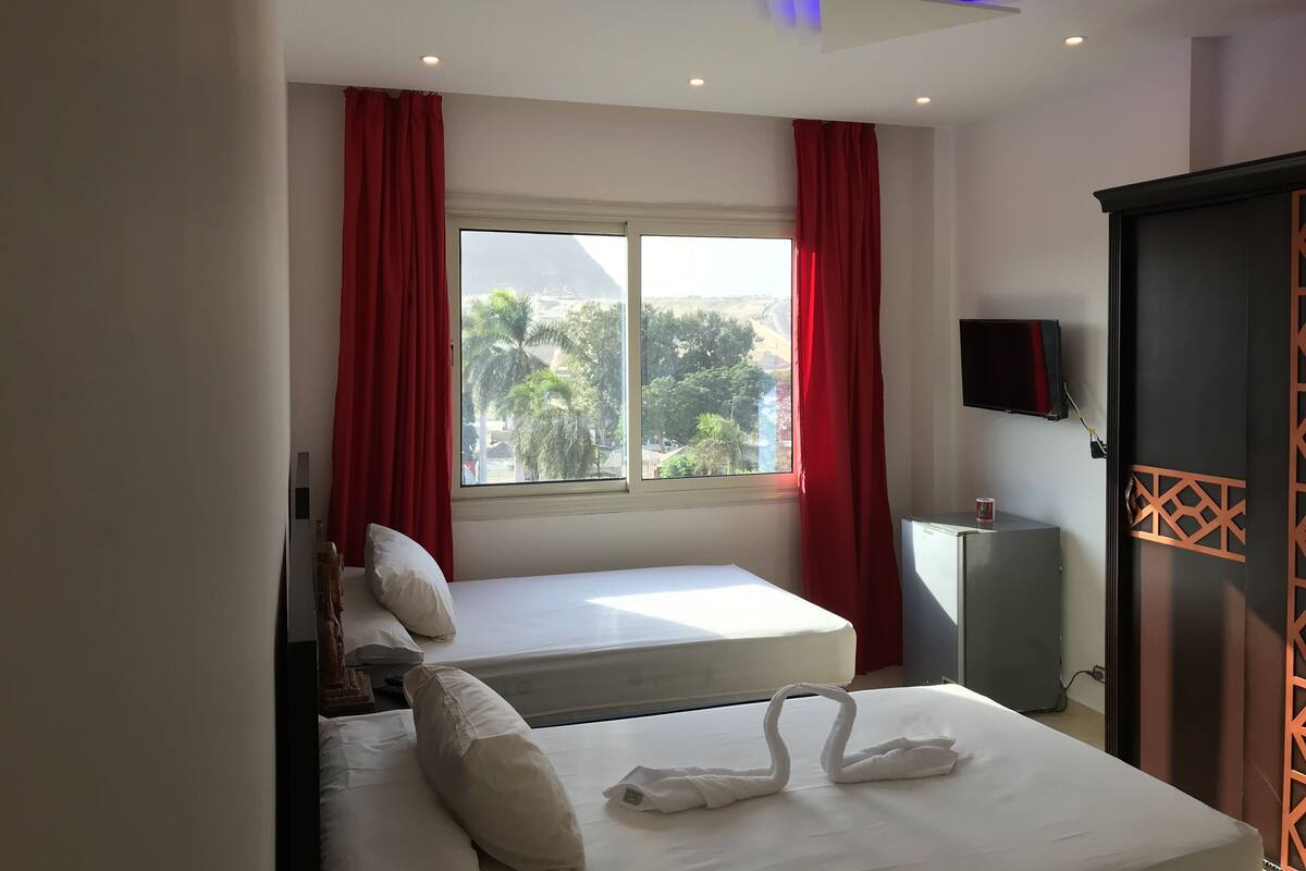 A hotel room with two beds, a TV, a desk, and a window with red curtains.