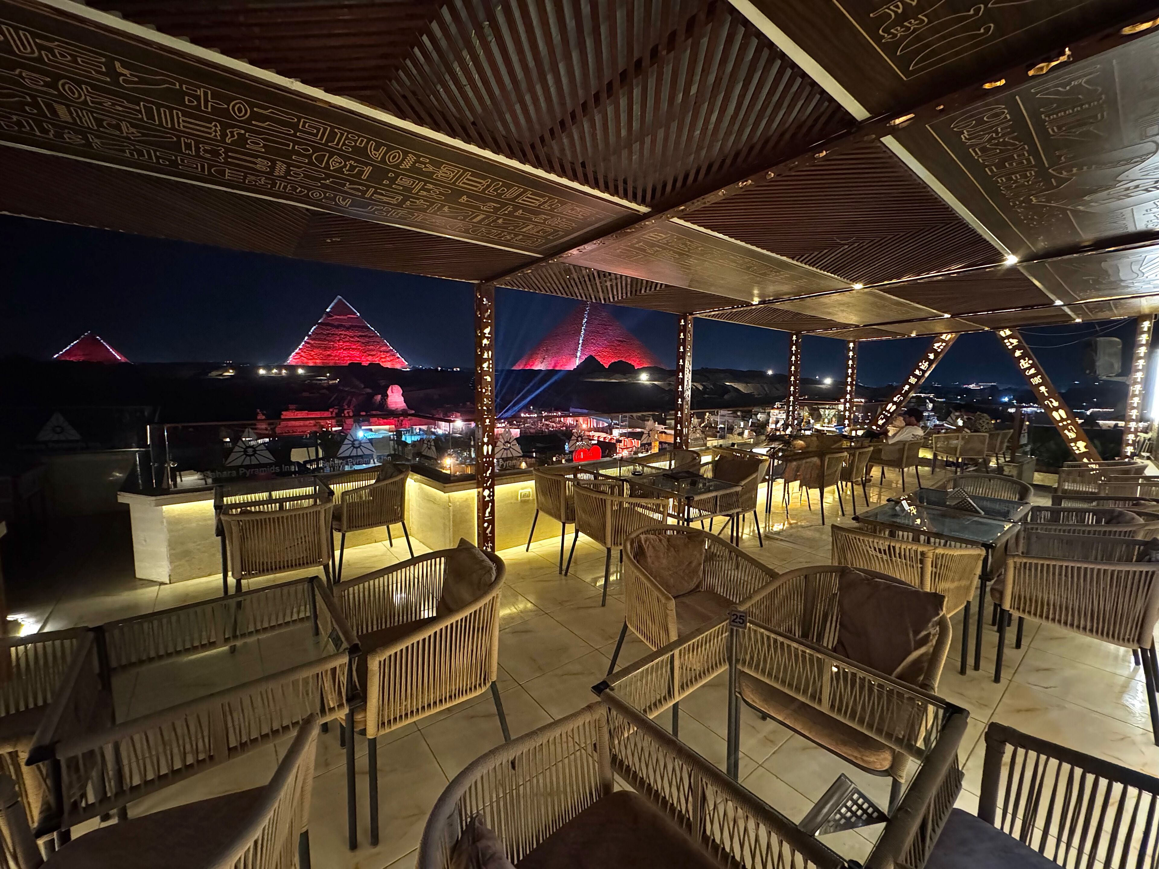 An outdoor seating area with wicker chairs and tables, a bar, and a view of a pyramid illuminated in red.
