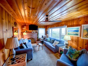 Cozy cabin interior with a seating area, fireplace, and large windows offering views of the surrounding landscape.