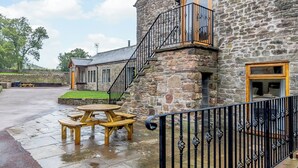A stone building with a staircase leading to a balcony, a wooden table and chairs outside, and a stone wall in the background. - The Old Granary - UK12135 (Hewelsfield, near Lydney)