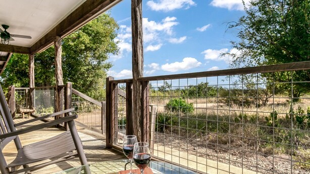 Una mesa de vidrio con dos copas de vino y un banco en un espacio exterior cubierto con vistas a un paisaje cercado.