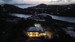 A residential area with a large house featuring a swimming pool, surrounded by hills and a body of water.