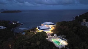 A coastal property with a swimming pool, a large house with a covered patio, and a view of the ocean.