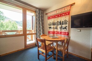 A cozy room with a wooden table and chairs, a large window with a view of greenery, a wall-mounted TV, and a patterned tapestry.