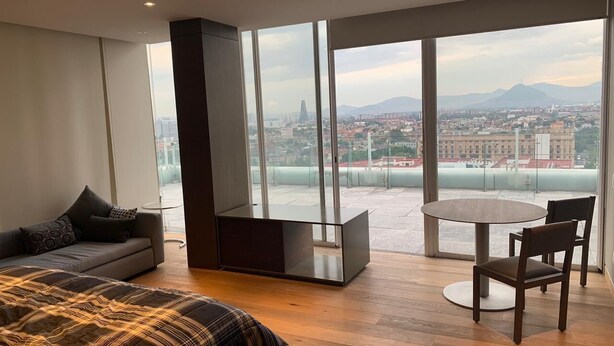 A modern hotel room with a large bed, a sofa, a dining table, and a view of a cityscape through large windows. - PH CIELO ALAMEDA, JUAREZ 52 (Centro)