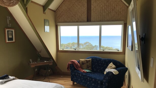 A bedroom with a large window, a bed, a blue patterned sofa, and a wooden desk. - Looking out over the ocean with uninterrupted views to Mayor Island and beyond! (Onemana)