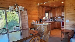 A wooden cabin interior with a dining area, kitchen, and a large window offering a view of greenery. - Enjoy the spectacular views from the balcony and cabin at this mountain location (Julian)