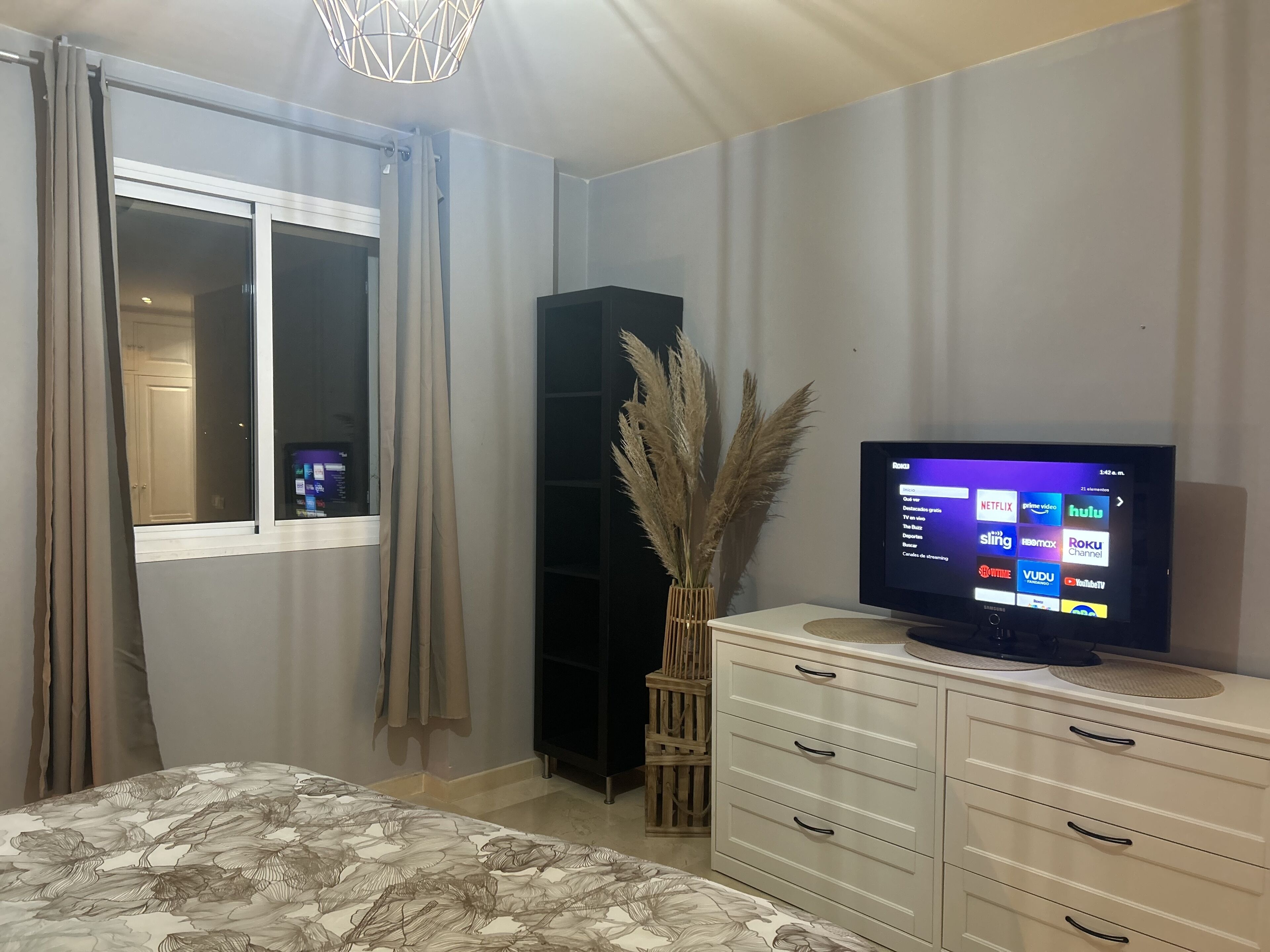 Een slaapkamer met een bed, een televisie op een standaard, een kledingkast en een raam met gordijnen.