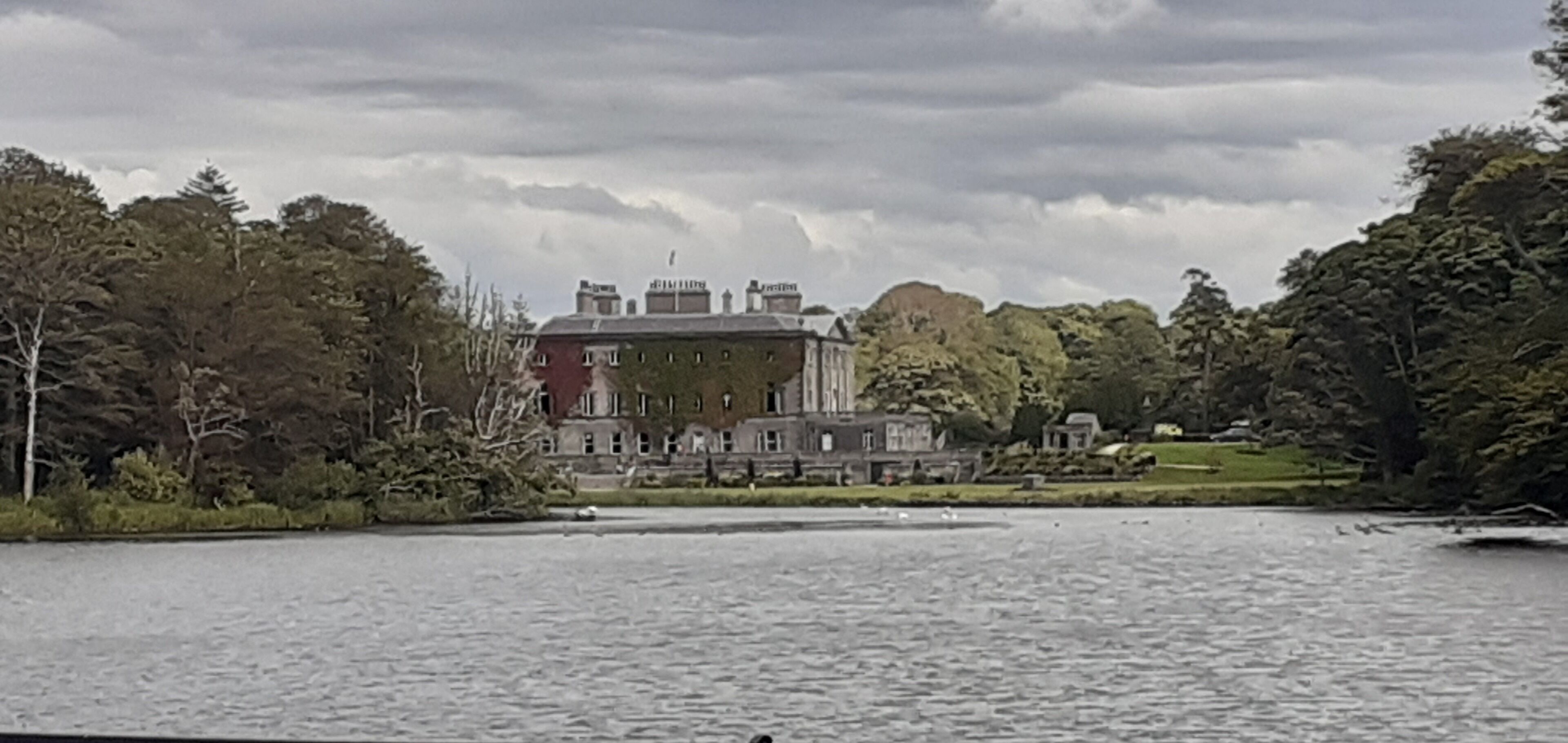 A large, historic building with a prominent tower, surrounded by lush greenery and a body of water.