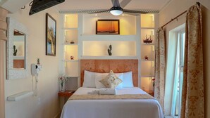 Una habitación de hotel con cama, cabecera, mesita de noche, espejo, teléfono en la pared, ventana con cortinas y ventilador de techo.
