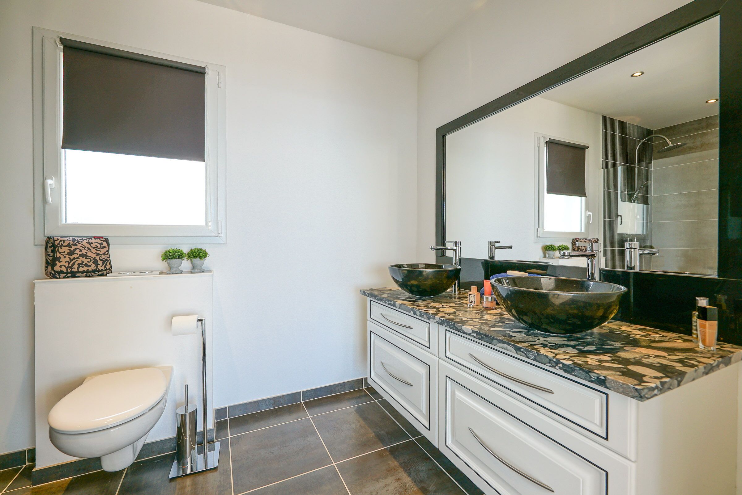 Une salle de bain moderne comprenant des toilettes, un bidet, deux lavabos avec de grands bacs, un grand miroir et une fenêtre avec un store.